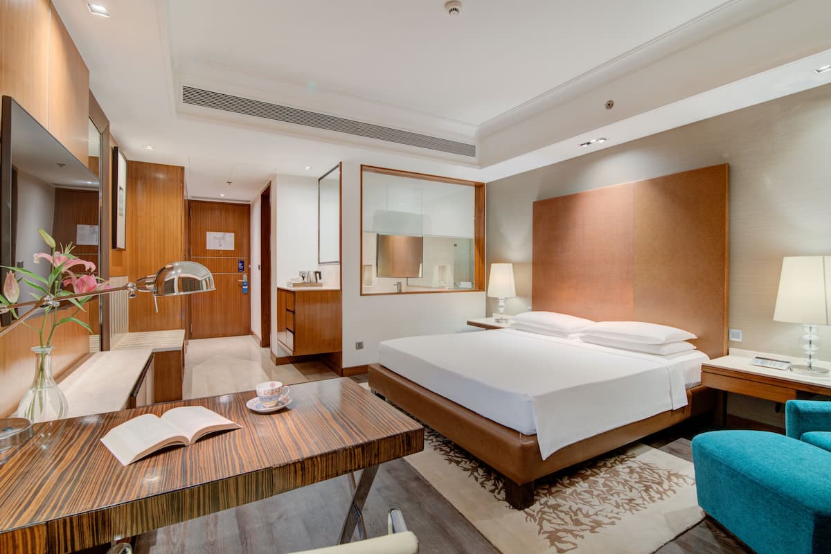 A modern hotel room with a large bed, a wooden desk, a chair, a mirror, and a bedside table with a lamp.