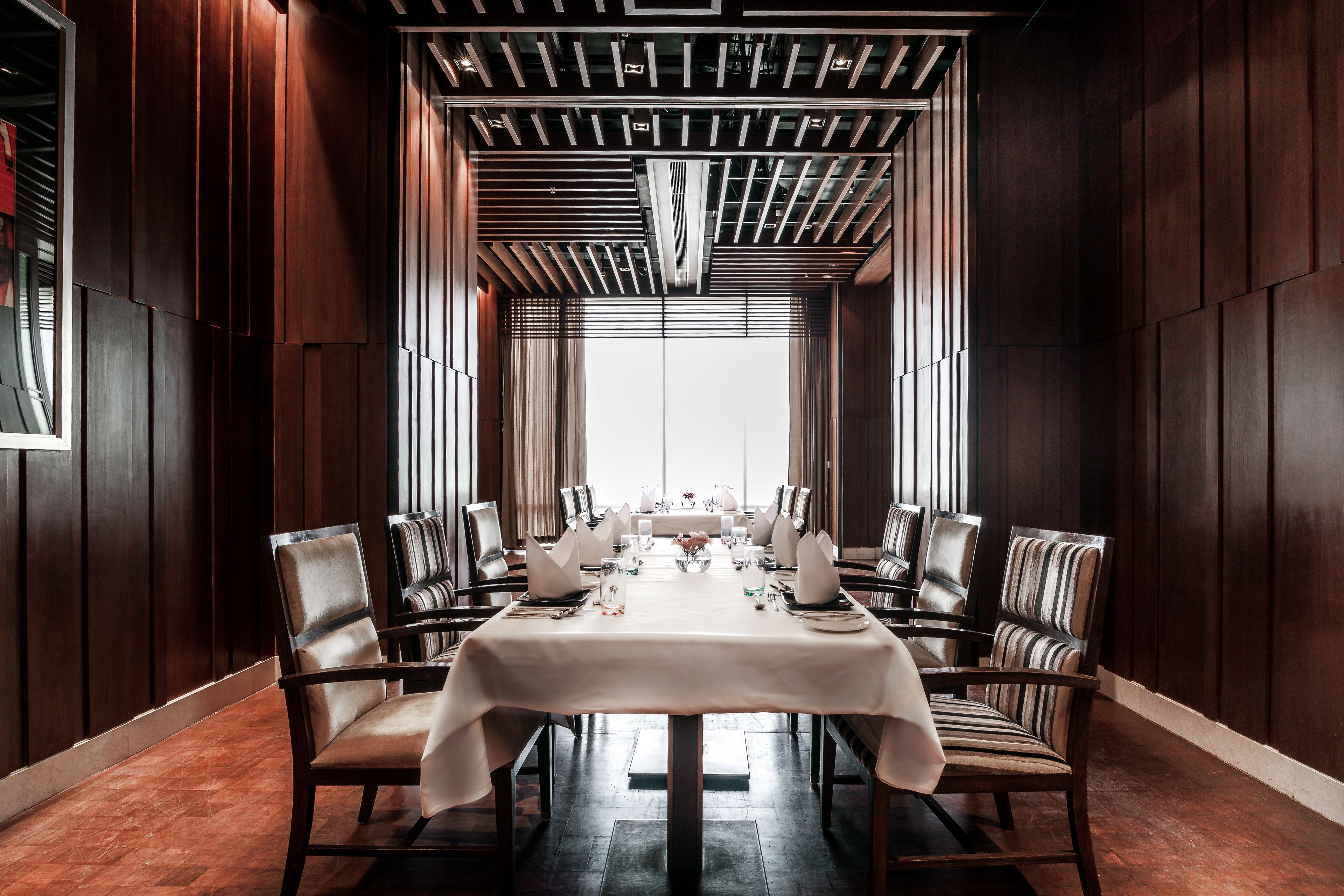A dining area with a long table set for a meal, surrounded by chairs, with a modern ceiling design and large windows in the background.