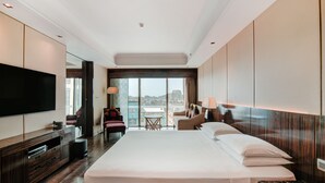 Una habitación de hotel moderna con una cama grande, un televisor de pantalla plana, una zona de estar con una mesa y sillas, y vistas a la ciudad.