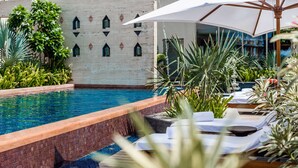Un área de piscina con sillas de descanso y sombrillas, rodeada de follaje y un muro decorativo.