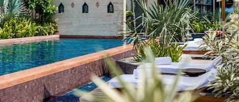 Un área de piscina con sillas de descanso y sombrillas, rodeada de follaje y un muro decorativo.