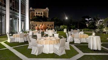 Un área de comedor al aire libre con mesas redondas y sillas blancas, frente a un gran edificio con arquitectura en forma de cúpula.