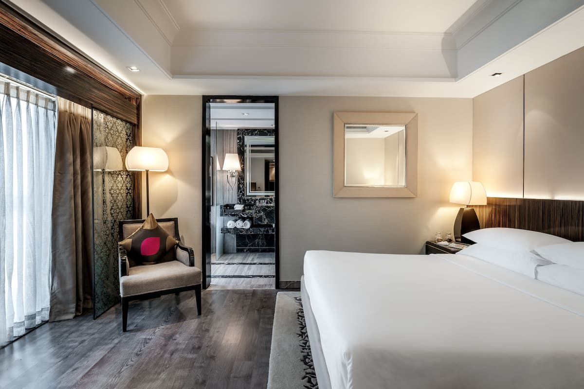 A hotel room with a large bed, a chair with a pillow, a nightstand with a lamp, a mirror, and a view of a bathroom with a sink and mirror.