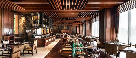 A spacious restaurant with wooden ceiling beams, a long counter with food displays, and large windows providing natural light.
