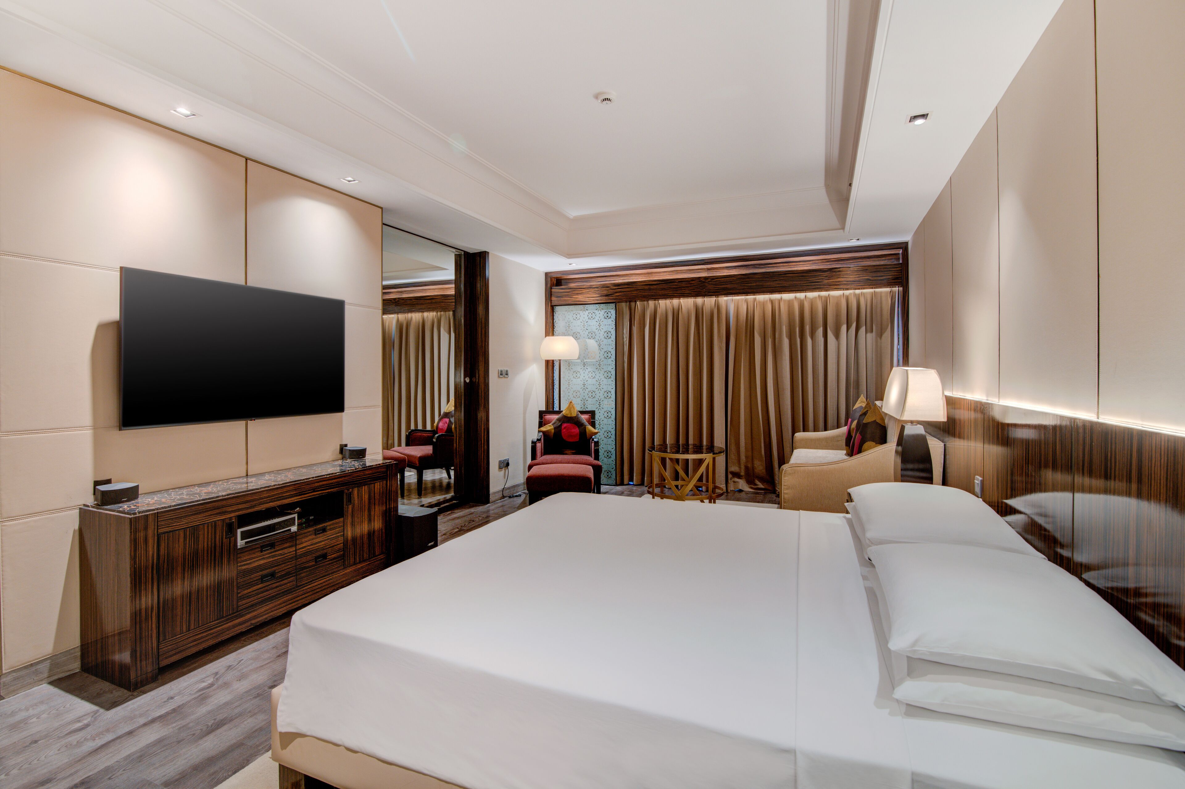 A modern hotel room with a large bed, a flat-screen TV mounted on the wall, a wooden dresser, a seating area with a chair and a small table, and a large window with curtains.