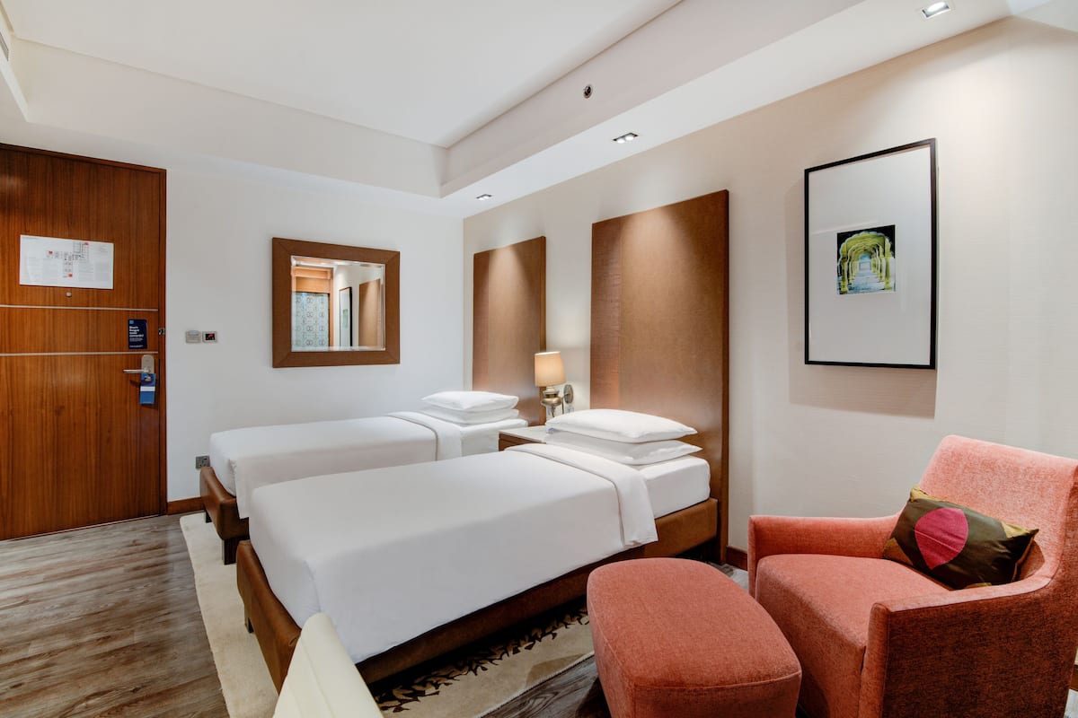 A hotel room with two beds, a wooden wardrobe, a mirror, a framed picture, and a pink armchair.