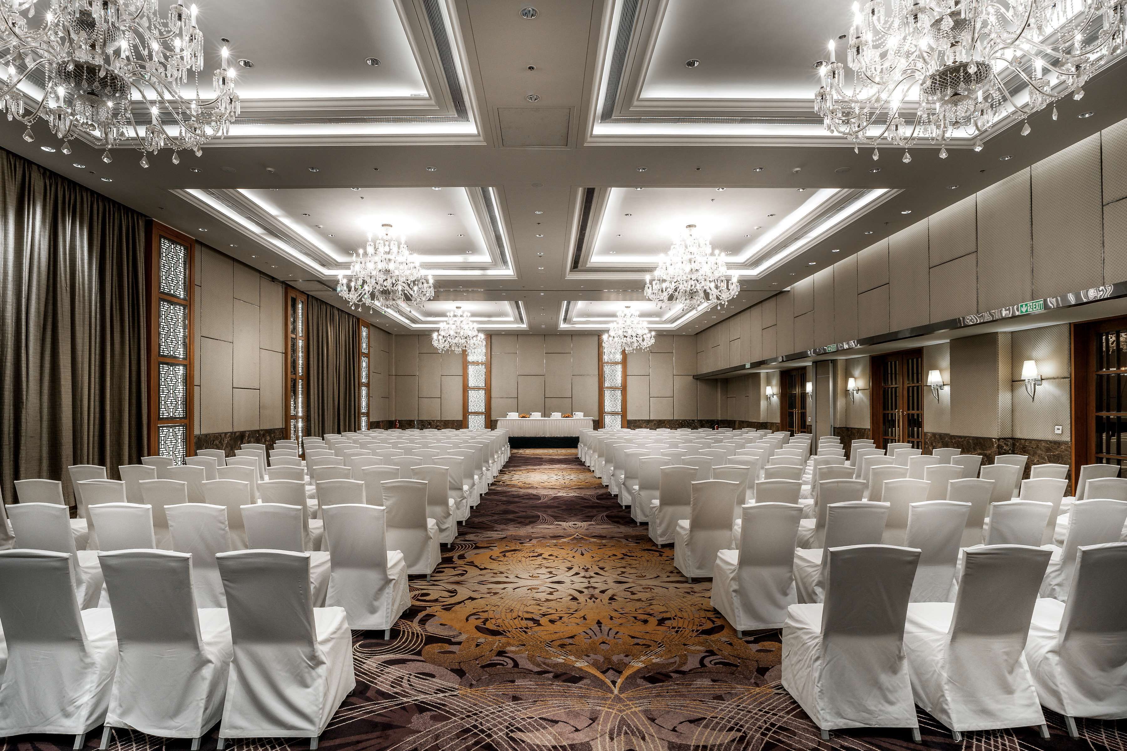 A conference room with rows of white chairs, chandeliers, and a carpeted floor.