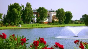A lake with a fountain, surrounded by lush greenery and a building with a red roof. - Hotel Isla Canela Golf (Ayamonte)