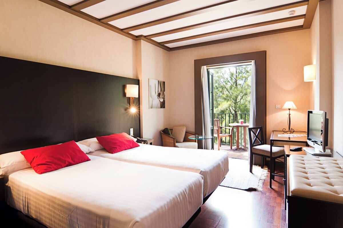 Habitación de hotel con dos camas, televisión, escritorio y balcón con vista a árboles.