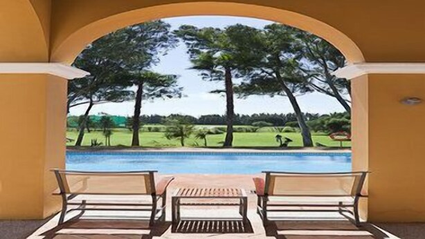 A poolside area with two chairs and a table, an arched doorway, and a view of a golf course. - Hotel Isla Canela Golf (Ayamonte)