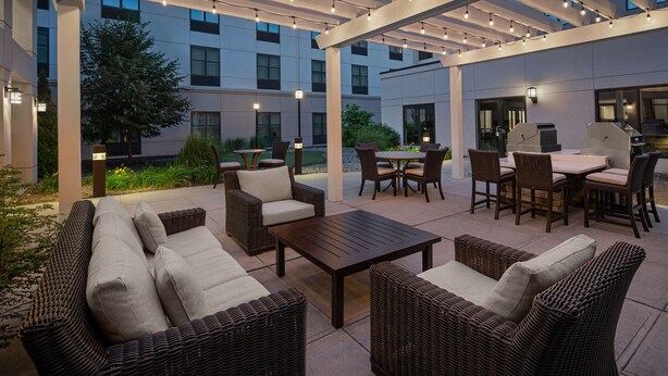 An outdoor patio area with wicker furniture, a wooden table, and a grill station. - Homewood Suites by Hilton Carle Place - Garden City, NY (Carle Place)
