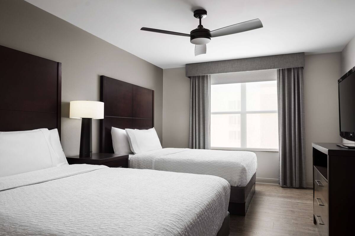 A hotel room with two beds, a ceiling fan, a TV, a nightstand with a lamp, and a window with curtains.