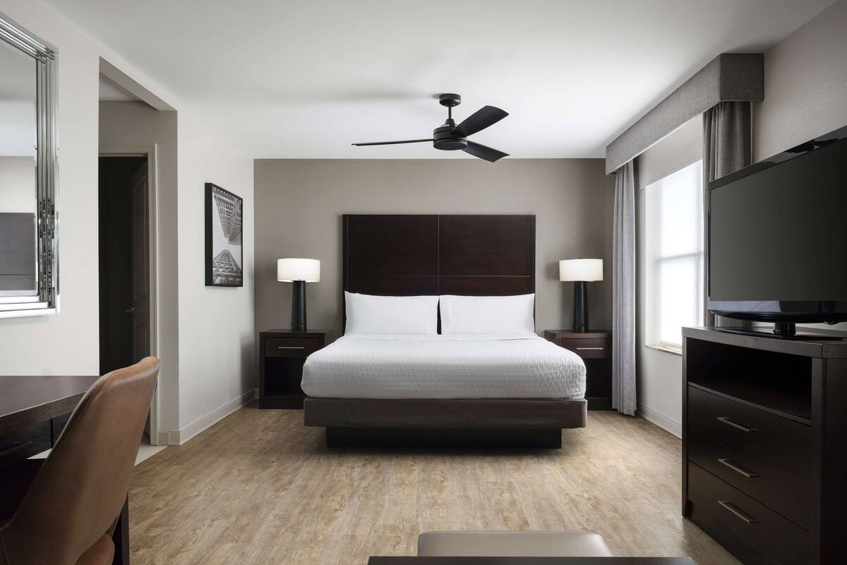 A hotel room with a bed, bedside tables with lamps, a ceiling fan, a TV on a cabinet, a desk, and a chair.