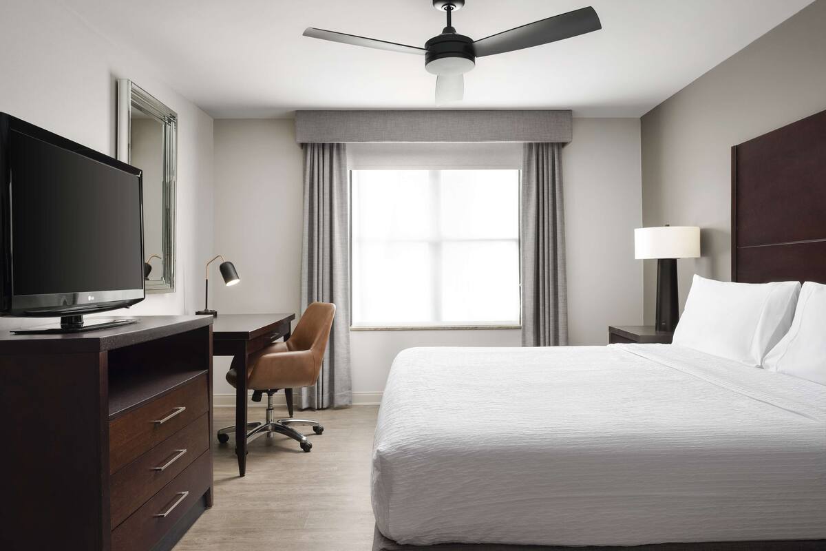 A hotel room with a bed, a desk with a computer, a chair, a TV, a ceiling fan, and a window with curtains.