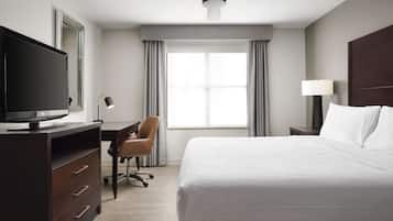 A hotel room with a bed, a desk with a computer, a chair, a TV, a ceiling fan, and a window with curtains.