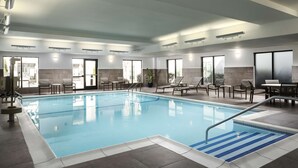 An indoor swimming pool with clear blue water, surrounded by lounge chairs and tables, and large windows allowing natural light. - Homewood Suites by Hilton Carle Place - Garden City, NY (Carle Place)