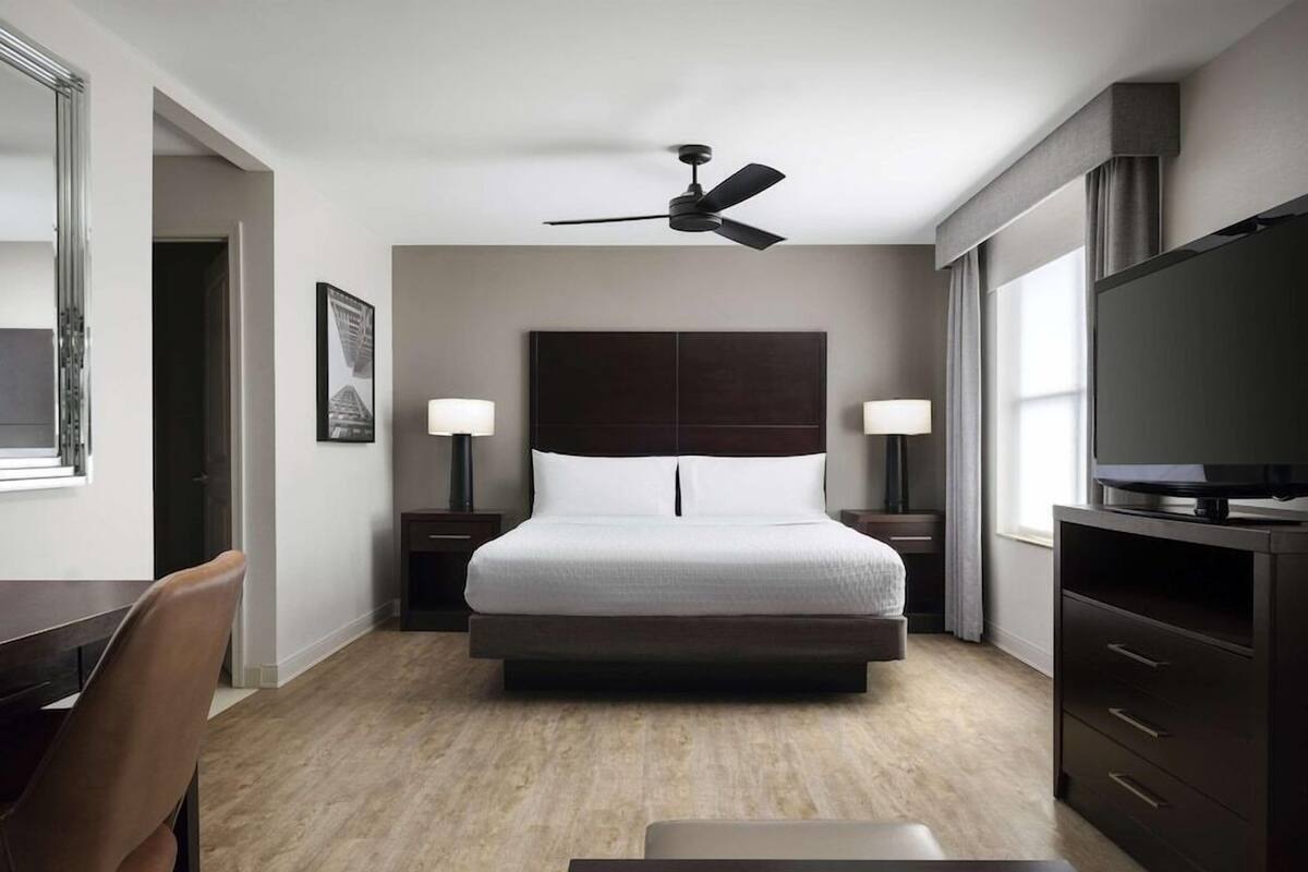 A hotel room with a large bed, a ceiling fan, a TV, a desk, and a chair.