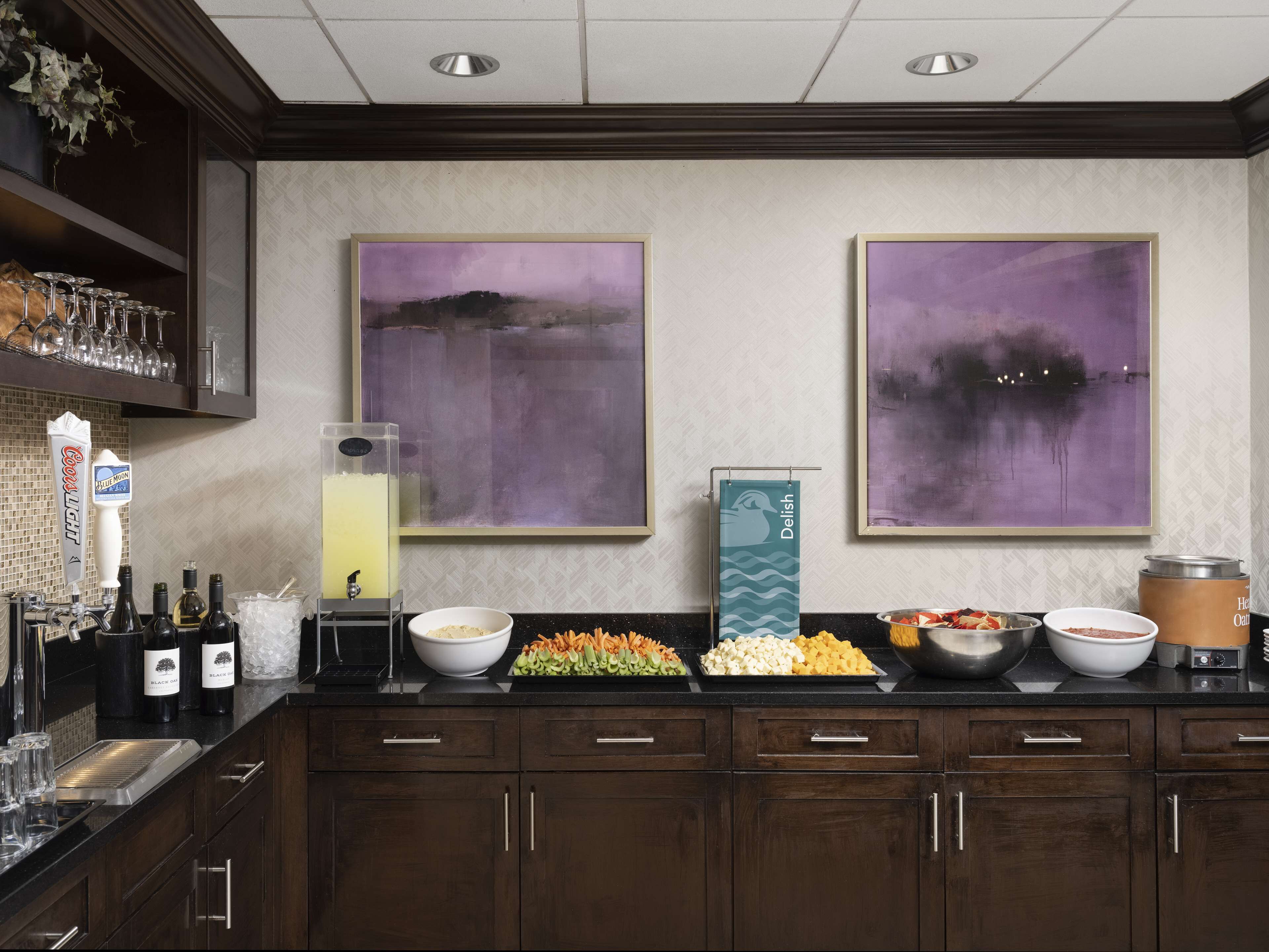 Angolo bar con distributore automatico di liquori, bicchieri da vino e diverse bottiglie. Due grandi dipinti astratti adornano la parete e un buffet offre diverse pietanze.