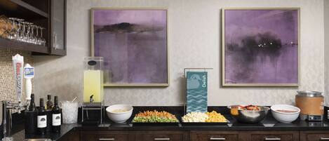 A bar area with a liquor dispenser, wine glasses, and a variety of bottles. There are two large abstract paintings on the wall and a buffet with bowls of food.