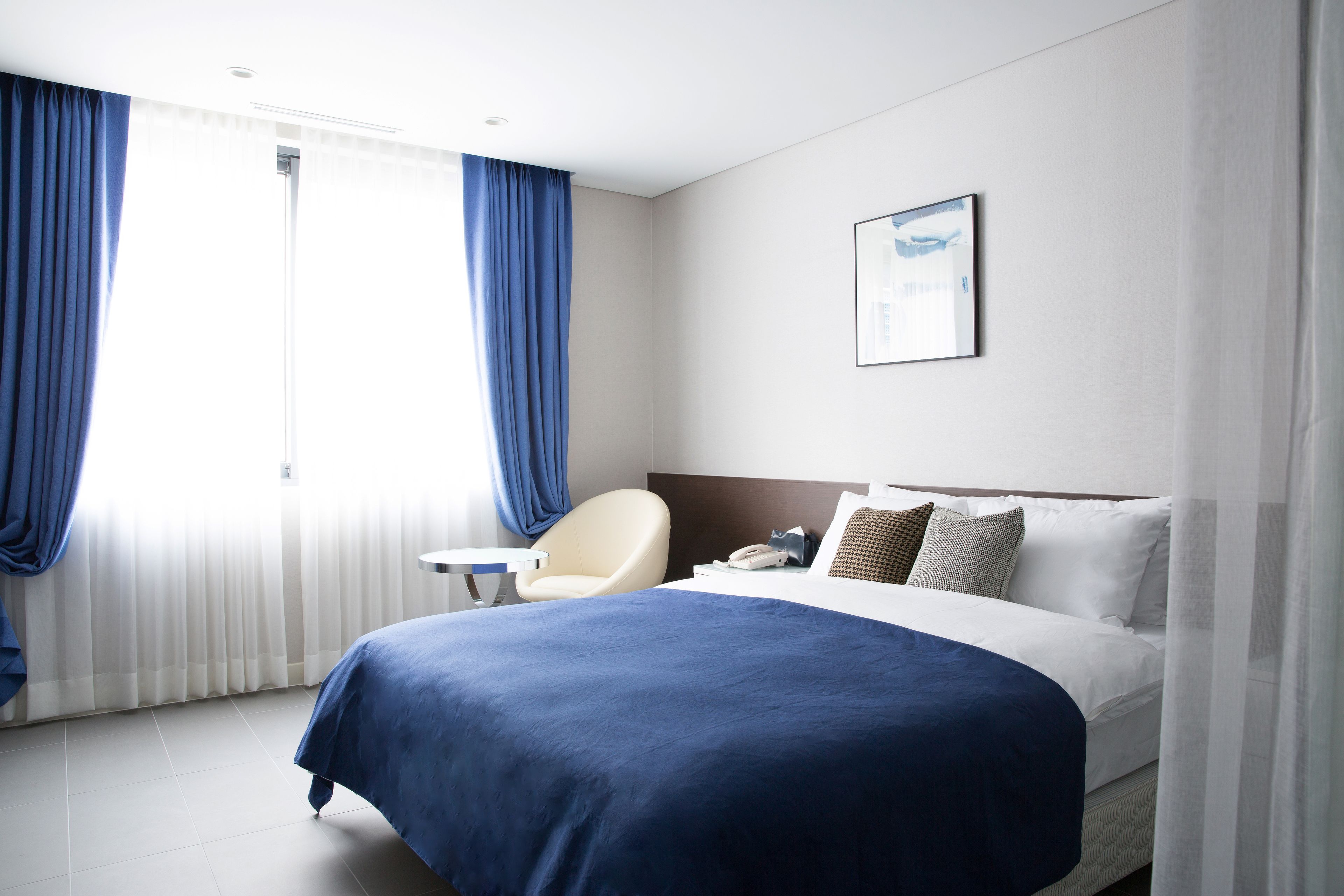 A hotel room with a large bed, a chair, a bedside table, and a window with curtains.