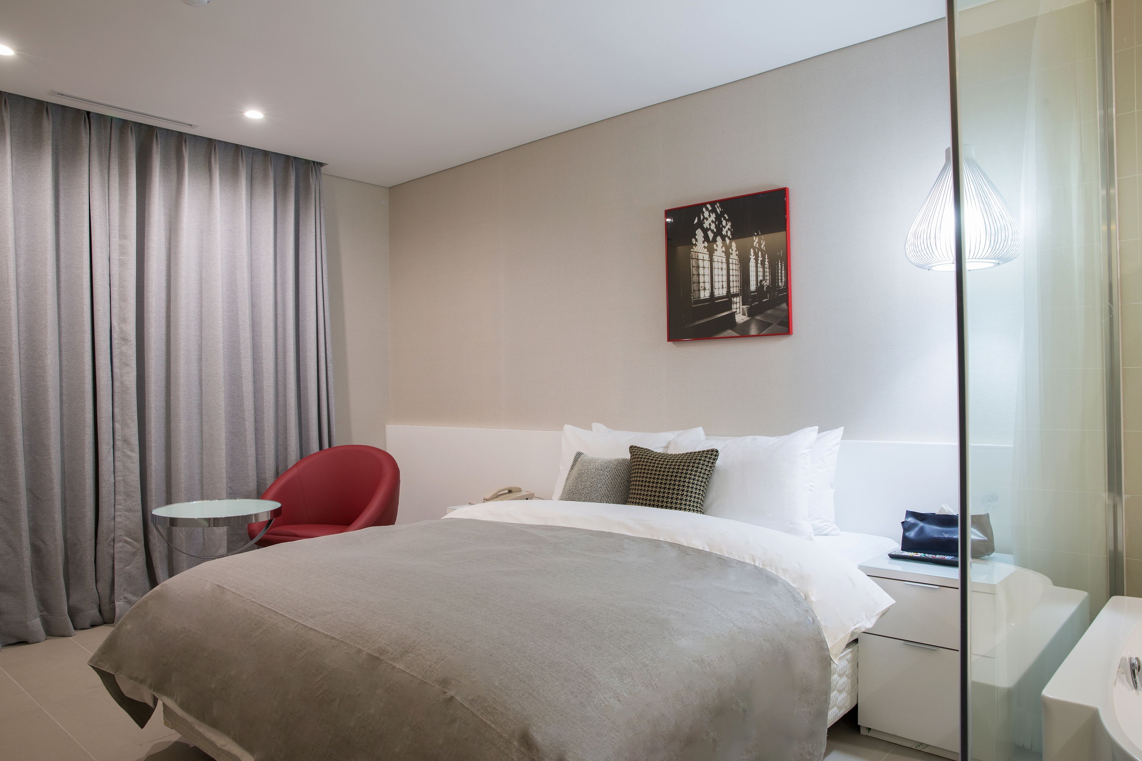 A modern hotel room with a large bed, a red chair, a small round table, a bedside table with a lamp, and a glass shower enclosure.