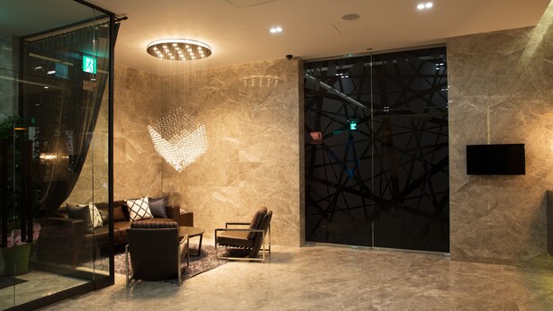Un hall d’entrée moderne avec un mur en verre, un espace détente avec un canapé et des fauteuils, un luminaire décoratif et un grand écran numérique.