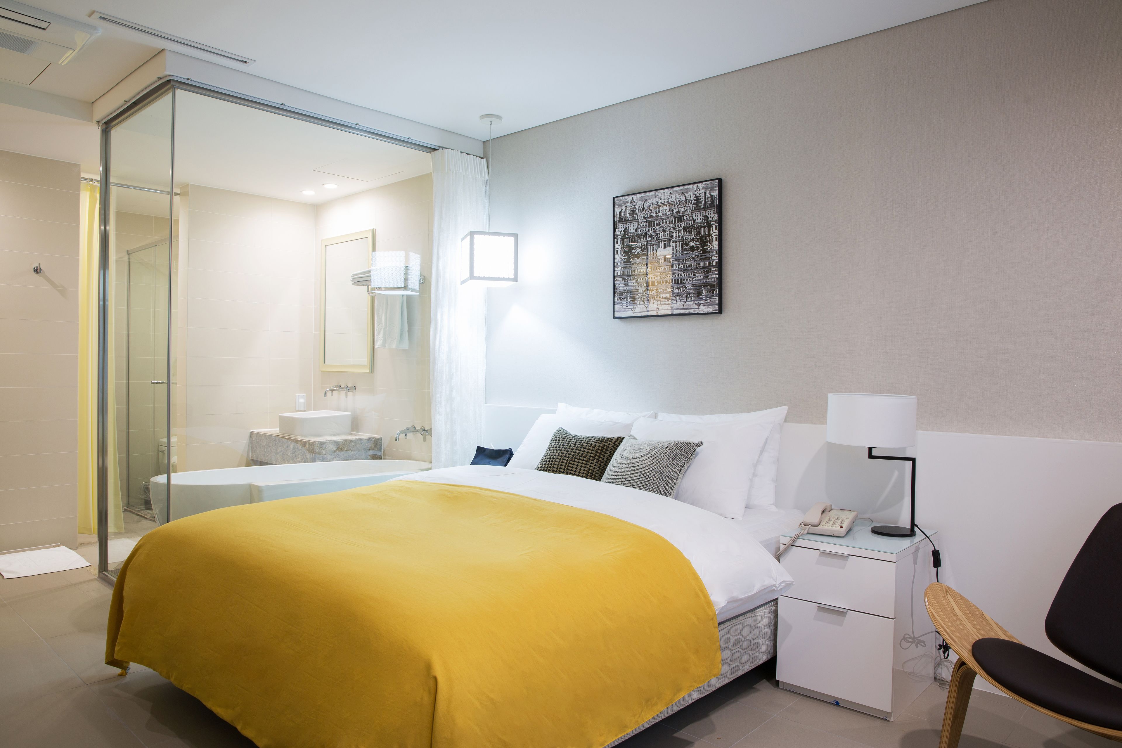 A modern hotel room with a large bed, a bedside table with a lamp, a chair, a bathroom with a glass shower enclosure, and a wall-mounted artwork.