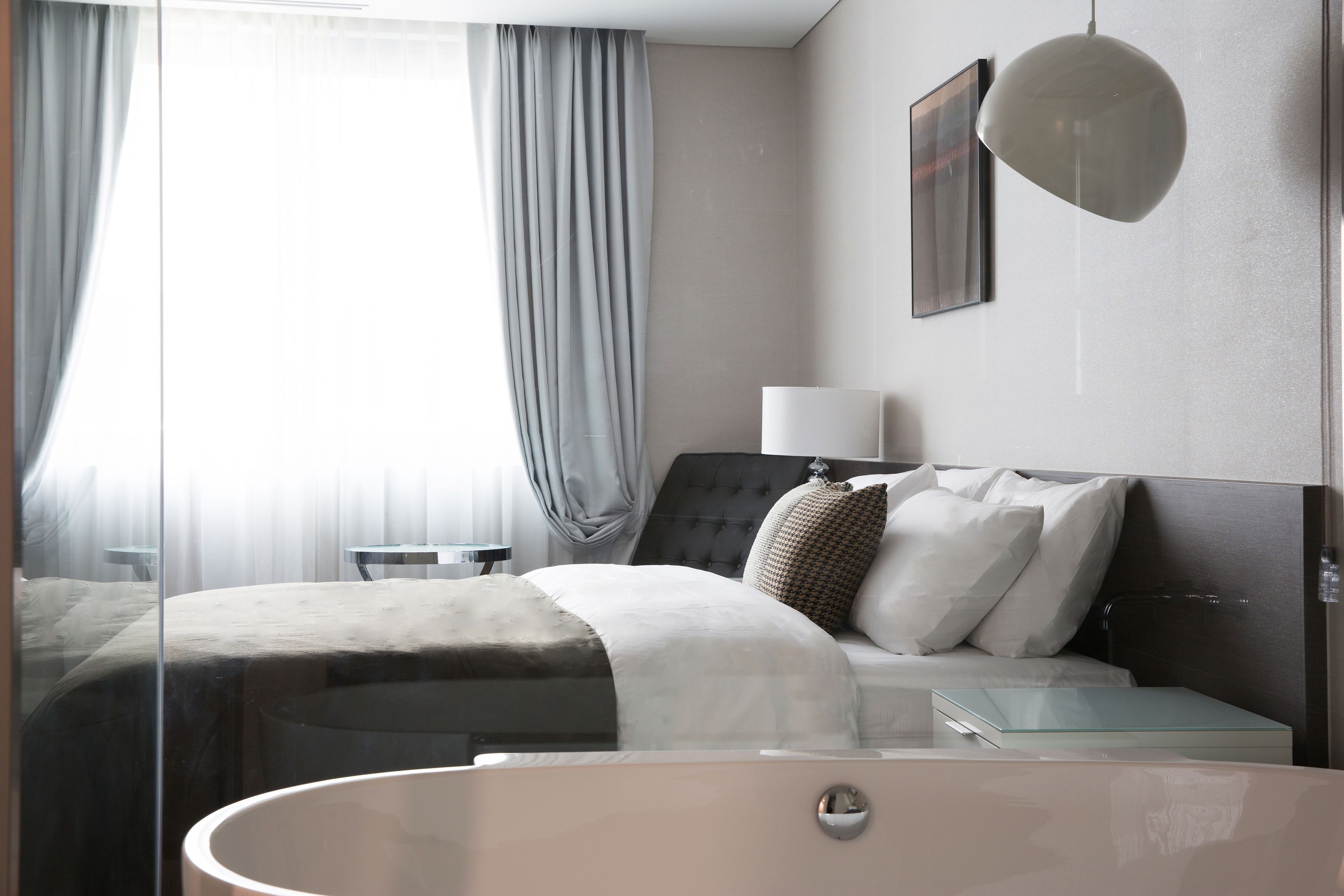 Una habitación de hotel moderna con una cama grande, una mesita de noche, una bañera de hidromasaje con mamparas de cristal y vistas al exterior a través de cortinas translúcidas.