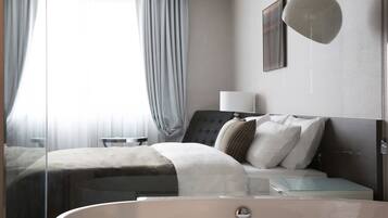 Una habitación de hotel moderna con una cama grande, una mesita de noche, una bañera de hidromasaje con mamparas de cristal y vistas al exterior a través de cortinas translúcidas.