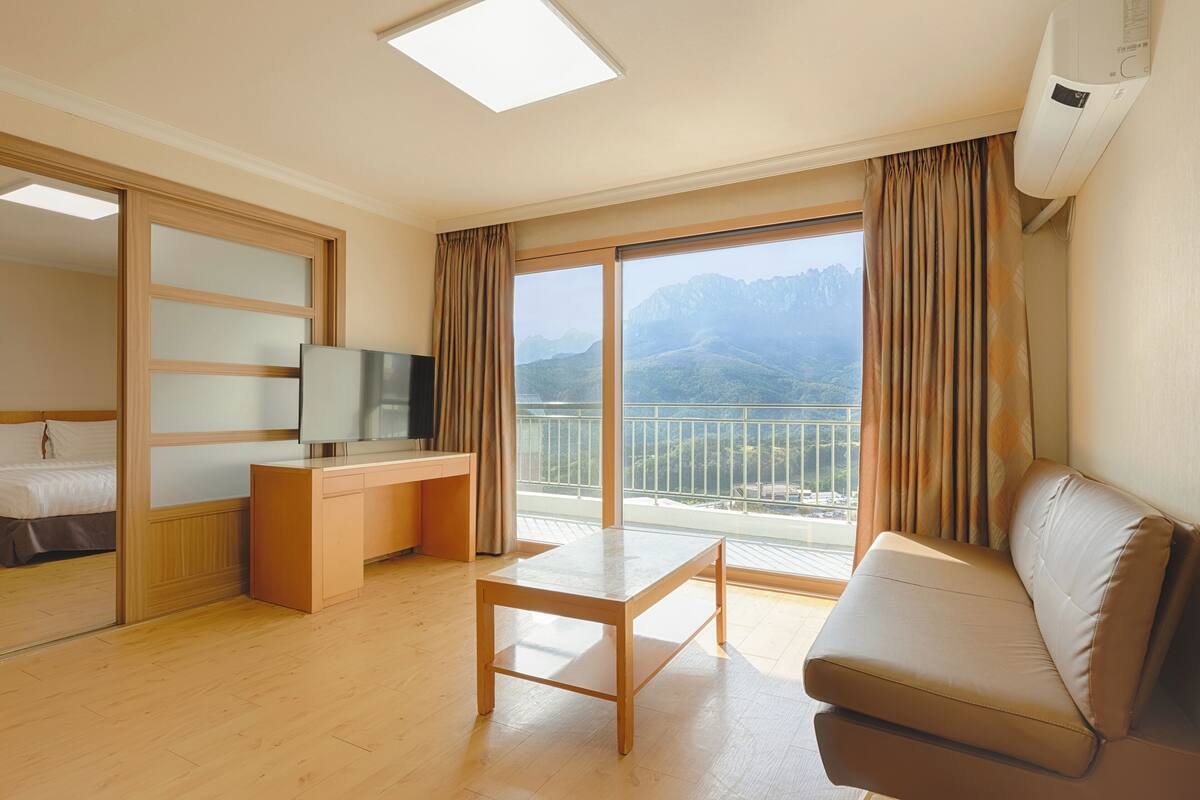 A hotel room with a bed, a desk, a TV, a sofa, and a balcony with a mountain view.