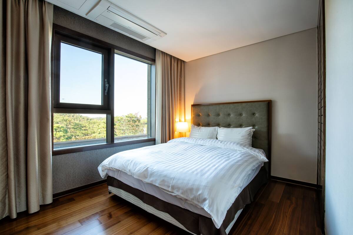 A hotel room with a large bed, a window with curtains, a wall-mounted air conditioner, and a nightstand with a lamp.