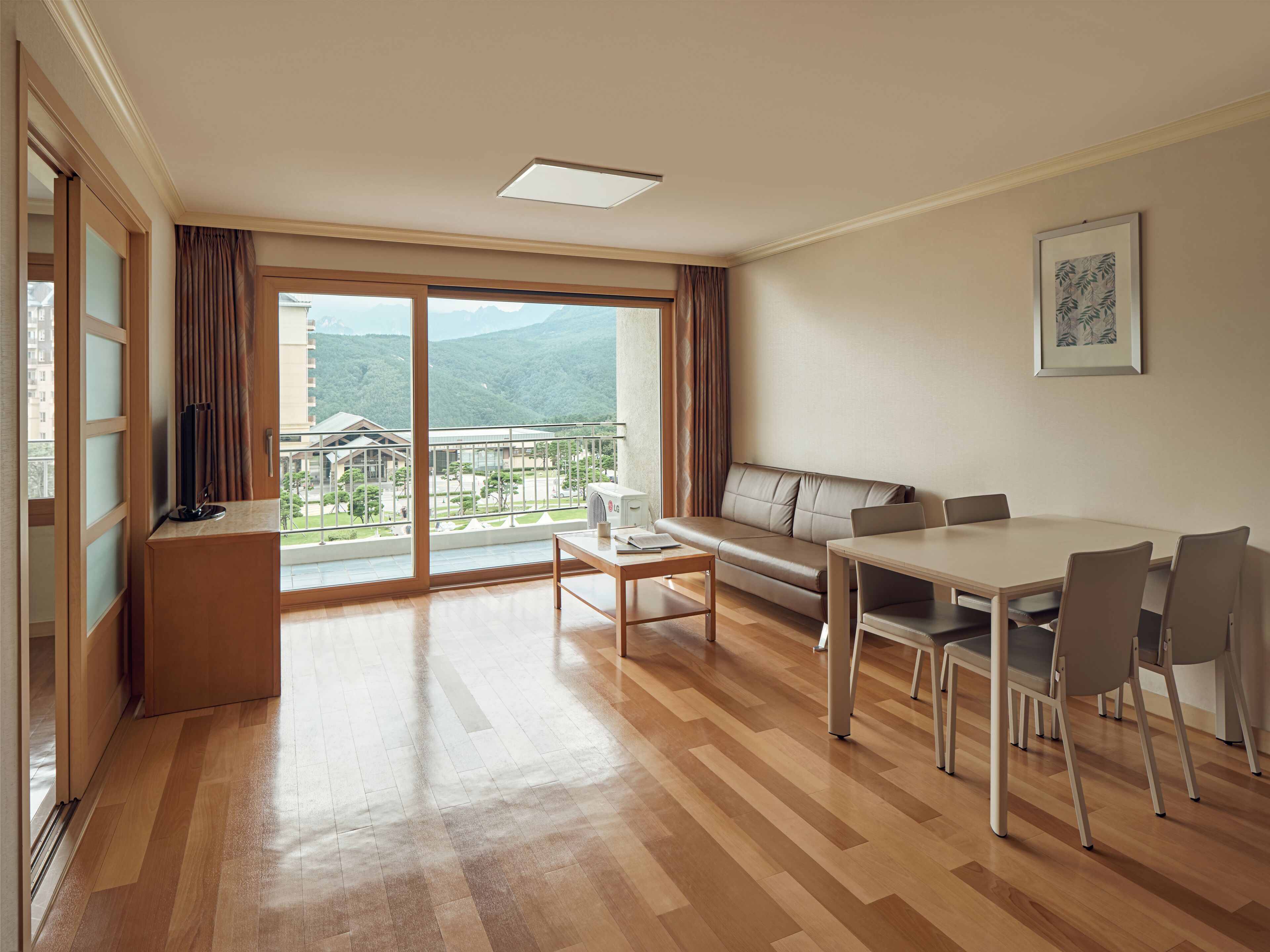 A modern hotel room with a wooden floor, a sofa, a dining table with chairs, and a large window offering a mountain view.
