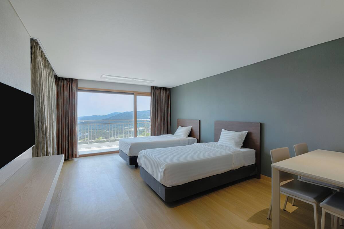 A hotel room with two beds, a dining table, and a large window offering a city view.