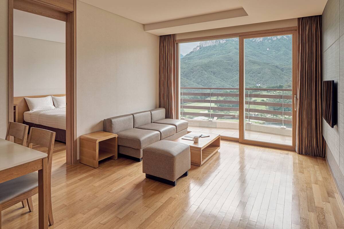 A modern hotel room with a wooden floor, a sofa, a bed, a dining table, and a large window offering a mountain view.