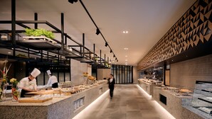 Un restaurant modern amb una barra llarga, cuiners preparant aliments i una paret amb un disseny decoratiu.