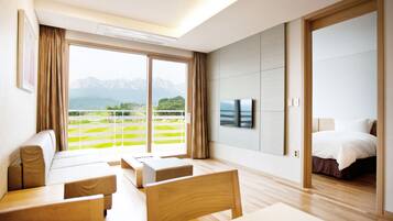 Una habitación de hotel moderna con un gran ventanal corredero que da a un balcón, una cama, un televisor y una zona de estar con vistas a las montañas.