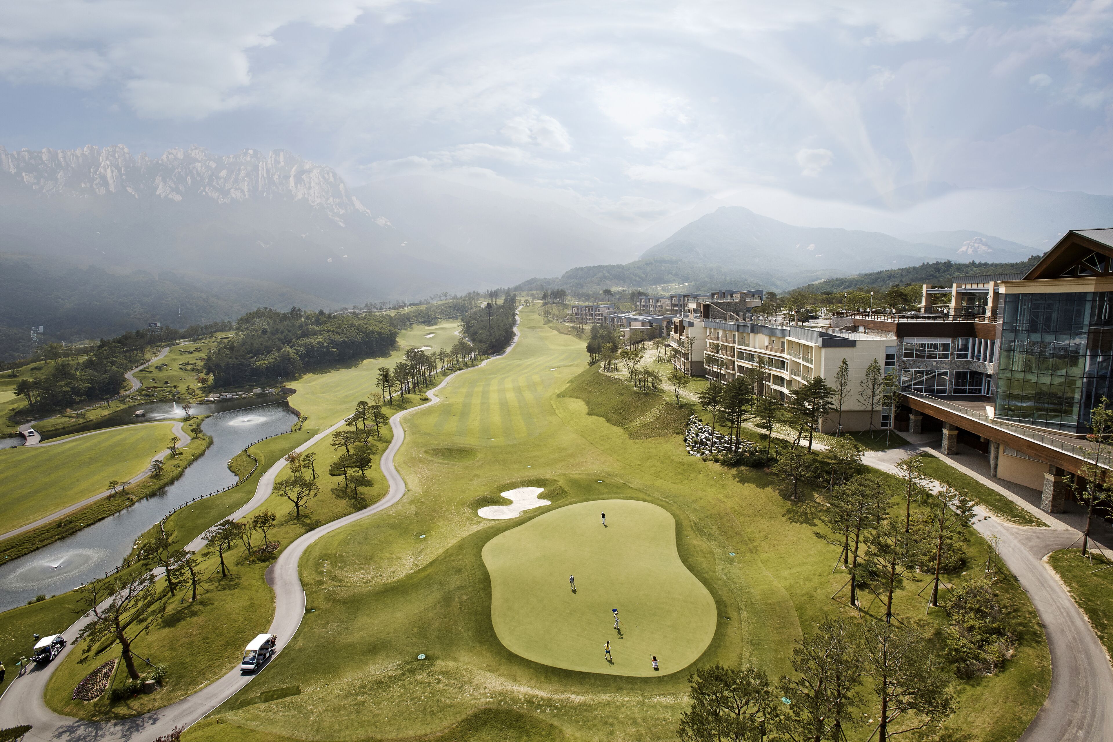 Campo de golf con vistas a montañas, un río y edificios modernos.