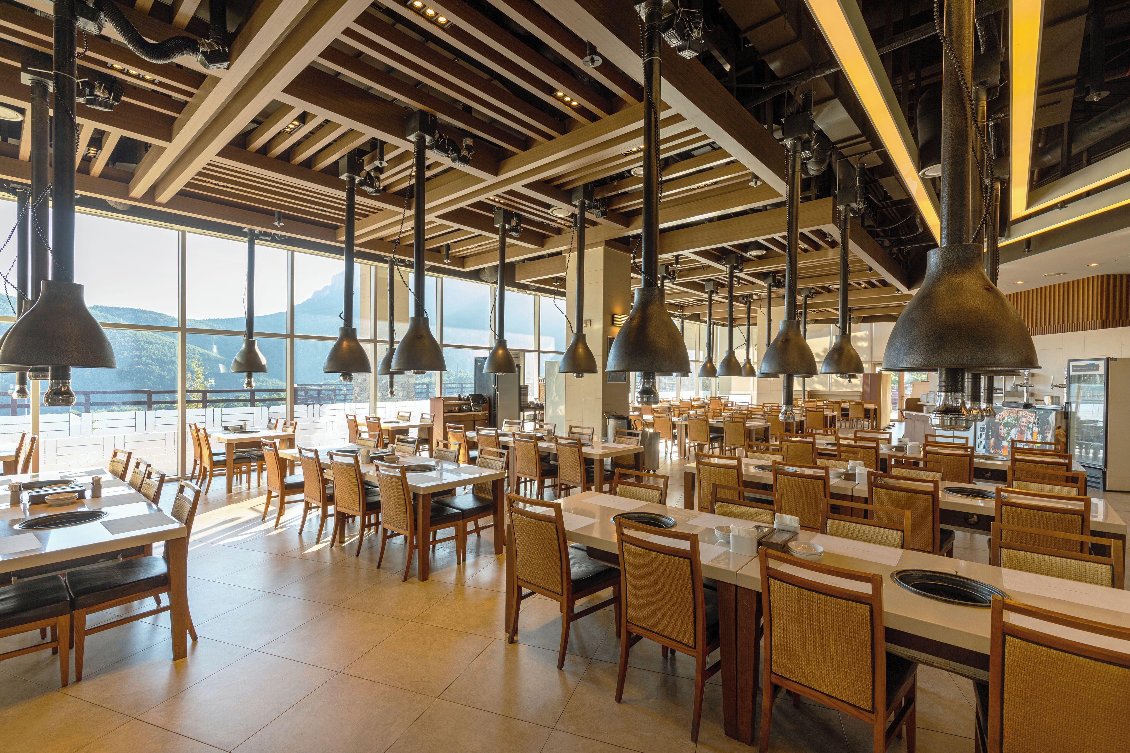 A spacious restaurant with high ceilings, large windows, and modern pendant lighting. Tables are arranged in a casual layout, and the interior features a mix of wood and metal elements.