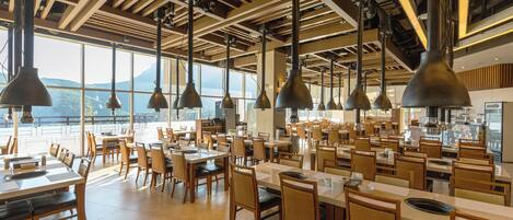A spacious restaurant with high ceilings, large windows, and modern pendant lighting. Tables are arranged in a casual layout, and the interior features a mix of wood and metal elements.