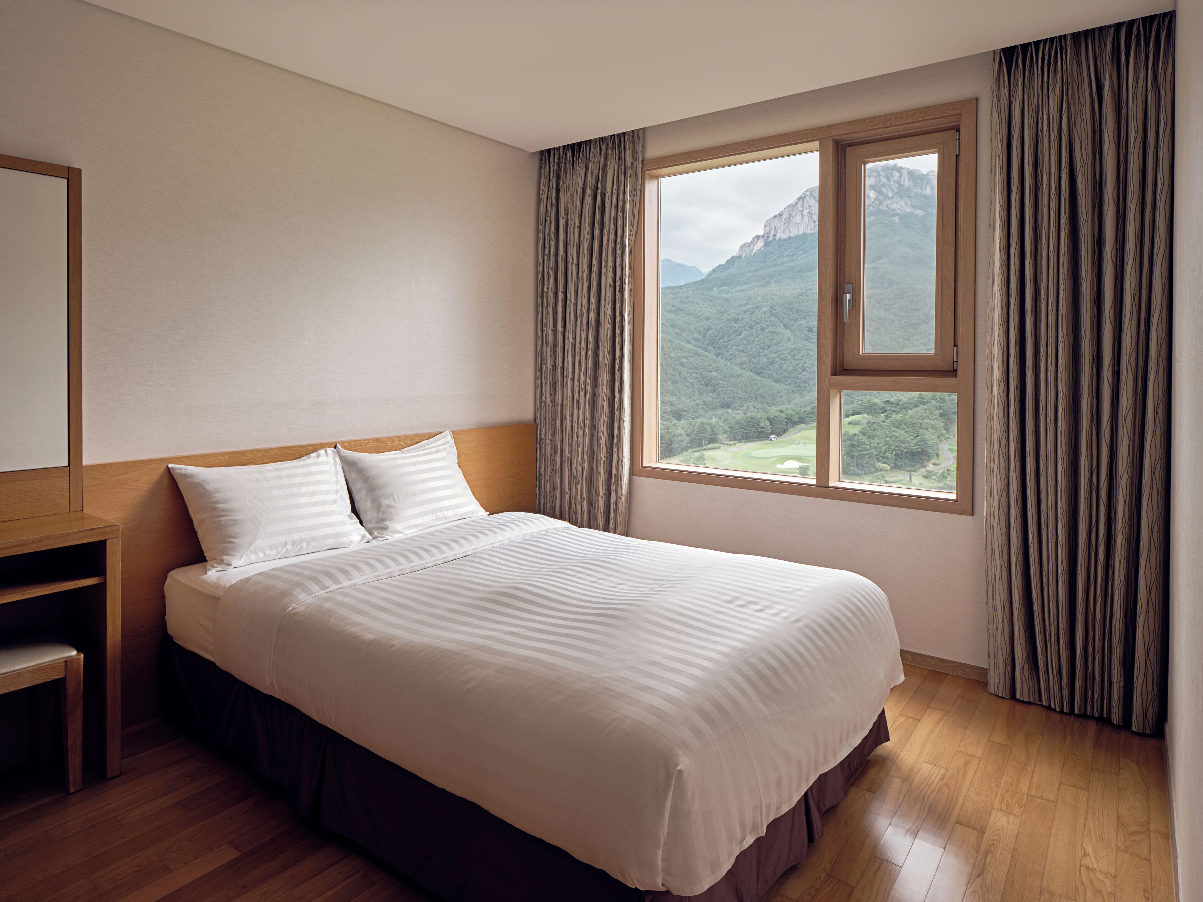 Habitación de hotel con una cama grande, una mesita de noche de madera y una ventana que ofrece vistas a un paisaje montañoso.