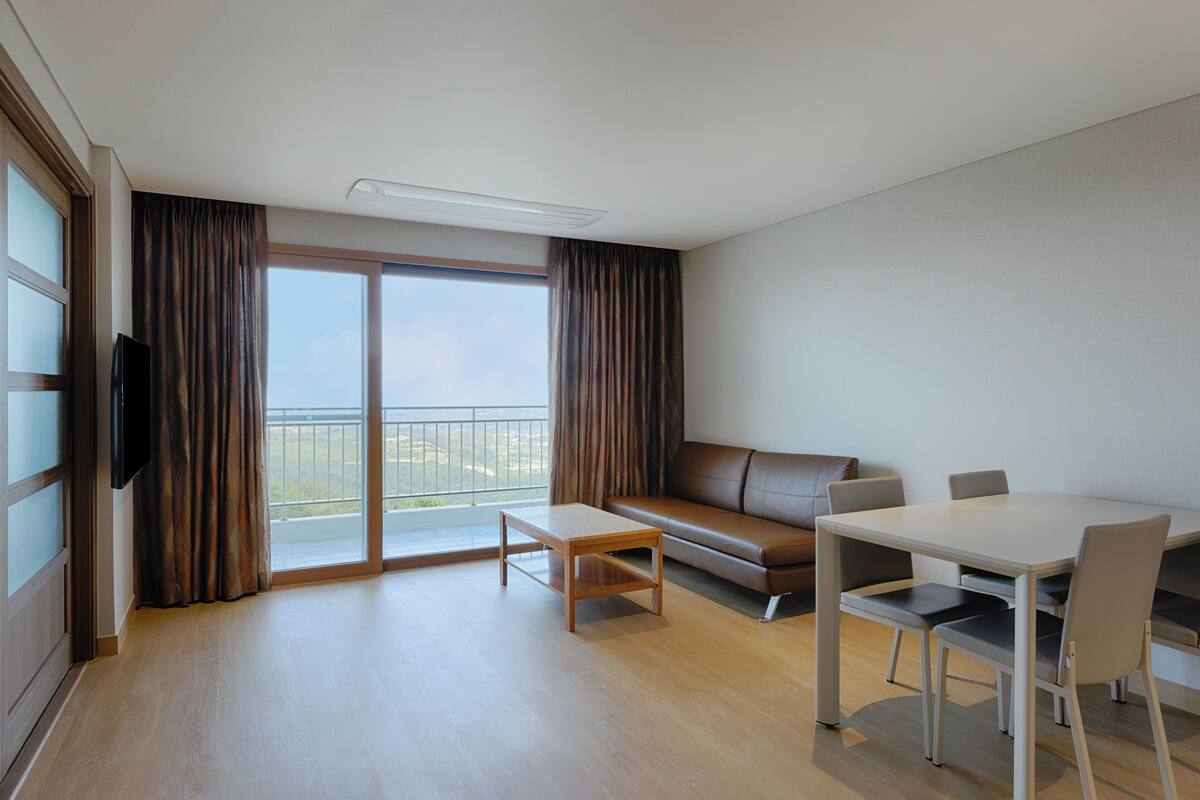 A modern hotel room with a sofa, a dining table with chairs, a flat-screen TV, and large windows with curtains.