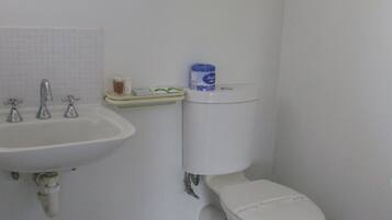 Un baño con lavamanos blanco, dos grifos, inodoro y un secamanos fijado a la pared.