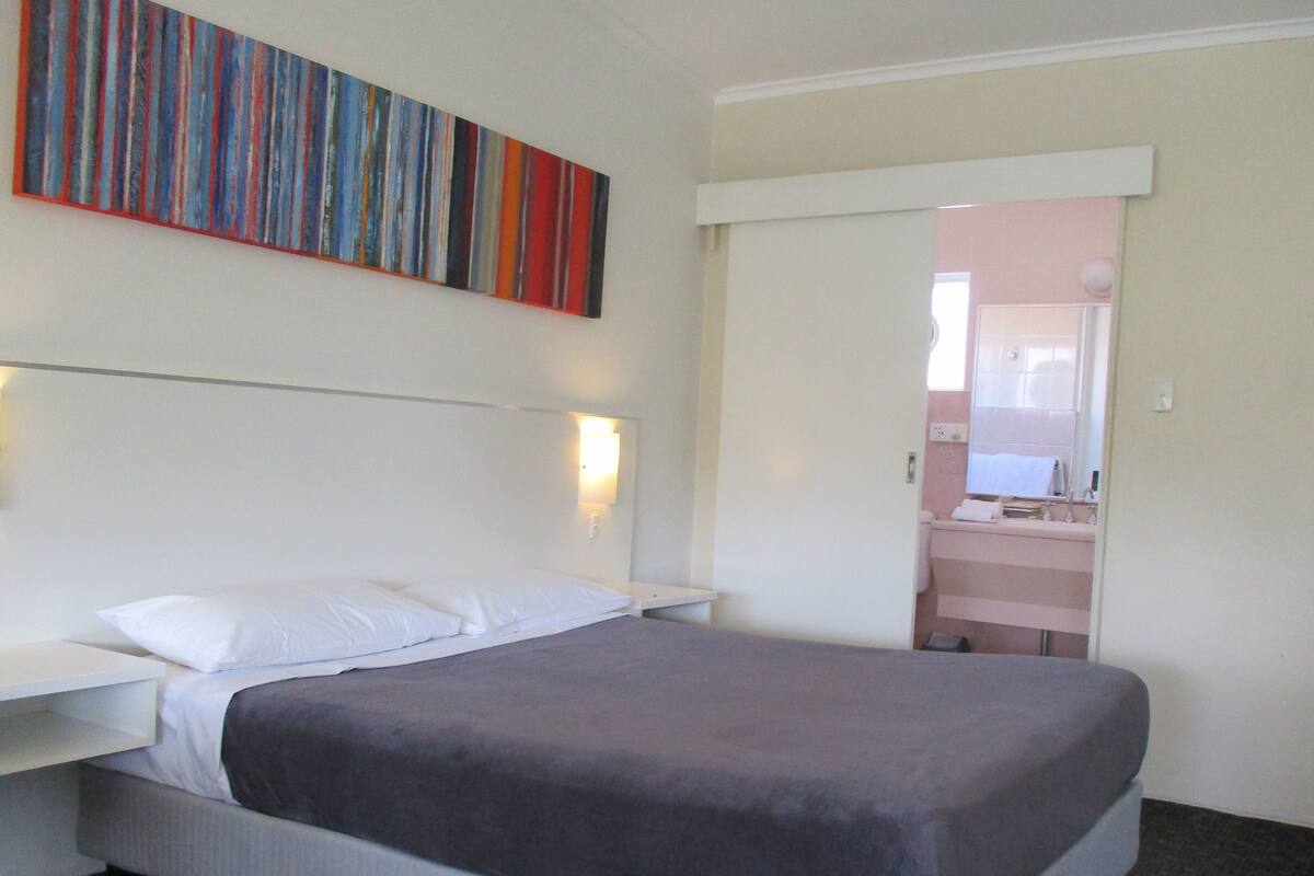 A hotel room with a bed, a nightstand, a wall-mounted light, and a bathroom visible through an open door.