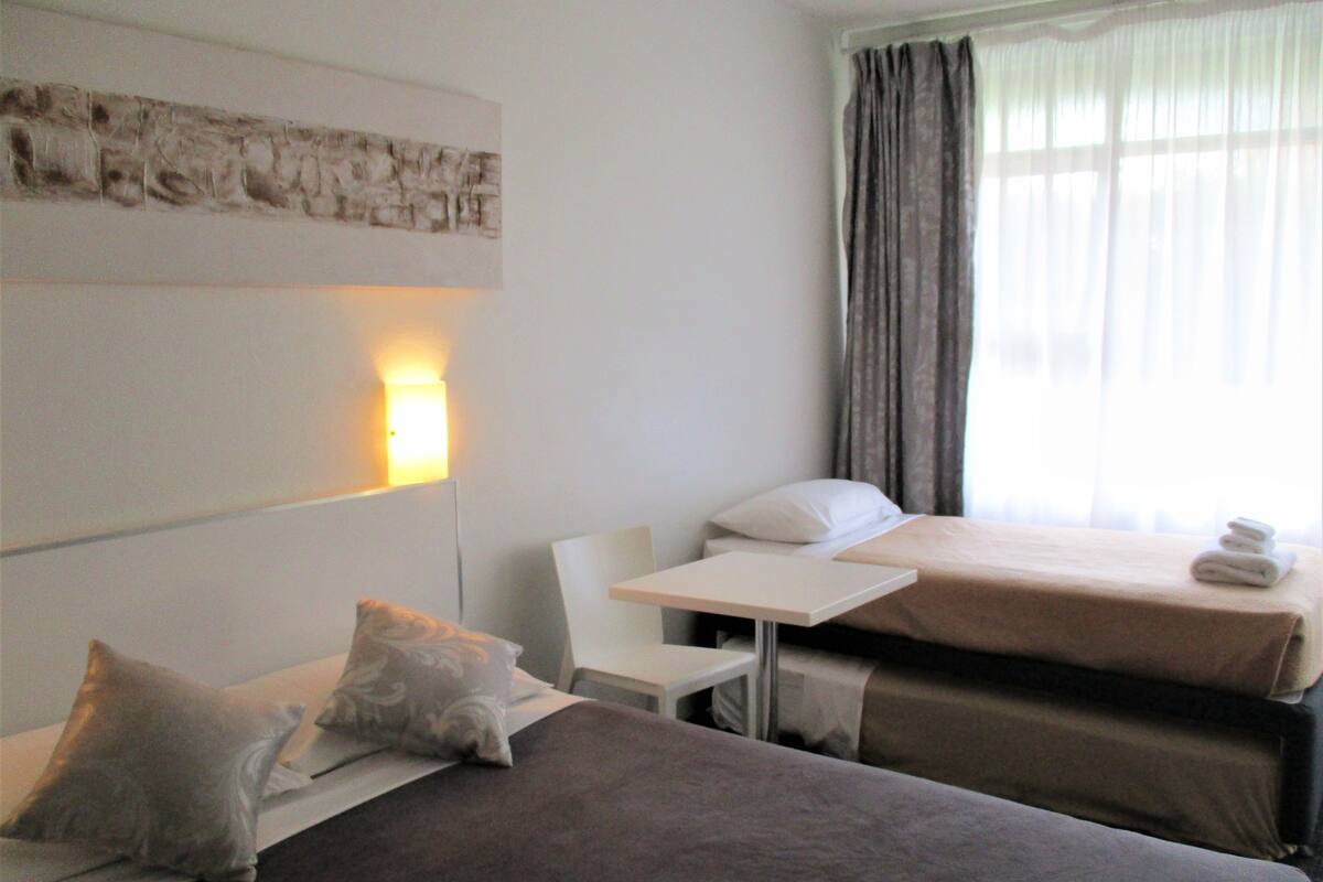 A hotel room with two beds, a small table, a chair, and a wall-mounted light.