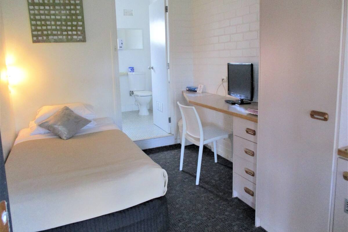 A small, compact room with a bed, a desk with a computer, and a chair. There is a door leading to a bathroom.