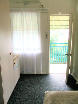 Une petite pièce, de taille modeste, comprenant un lit simple, une fenêtre avec des rideaux légers, un radiateur et donnant sur un balcon avec une rambarde verte.