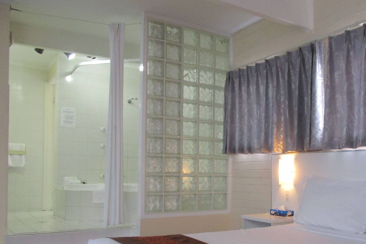 A hotel room with a bed, a shower area, and a window with curtains.