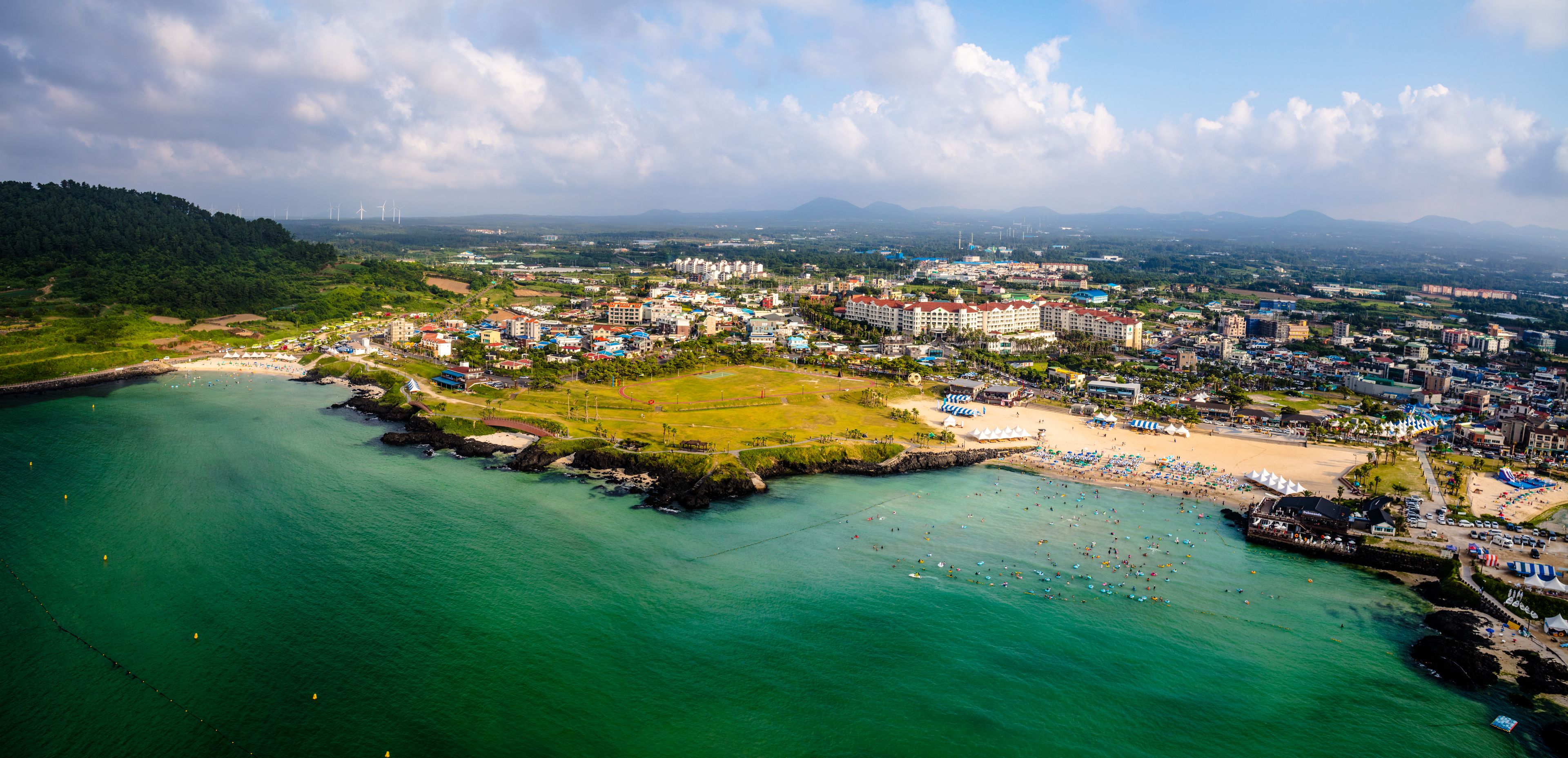 An aerial view of a coastal town with a beach, buildings, and a green park area.