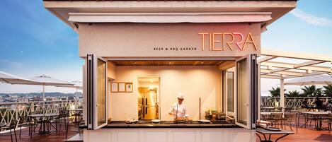 Quiosco de comida llamado "TERRA" con un cocinero preparando alimentos, rodeado de mesas y sillas al aire libre.