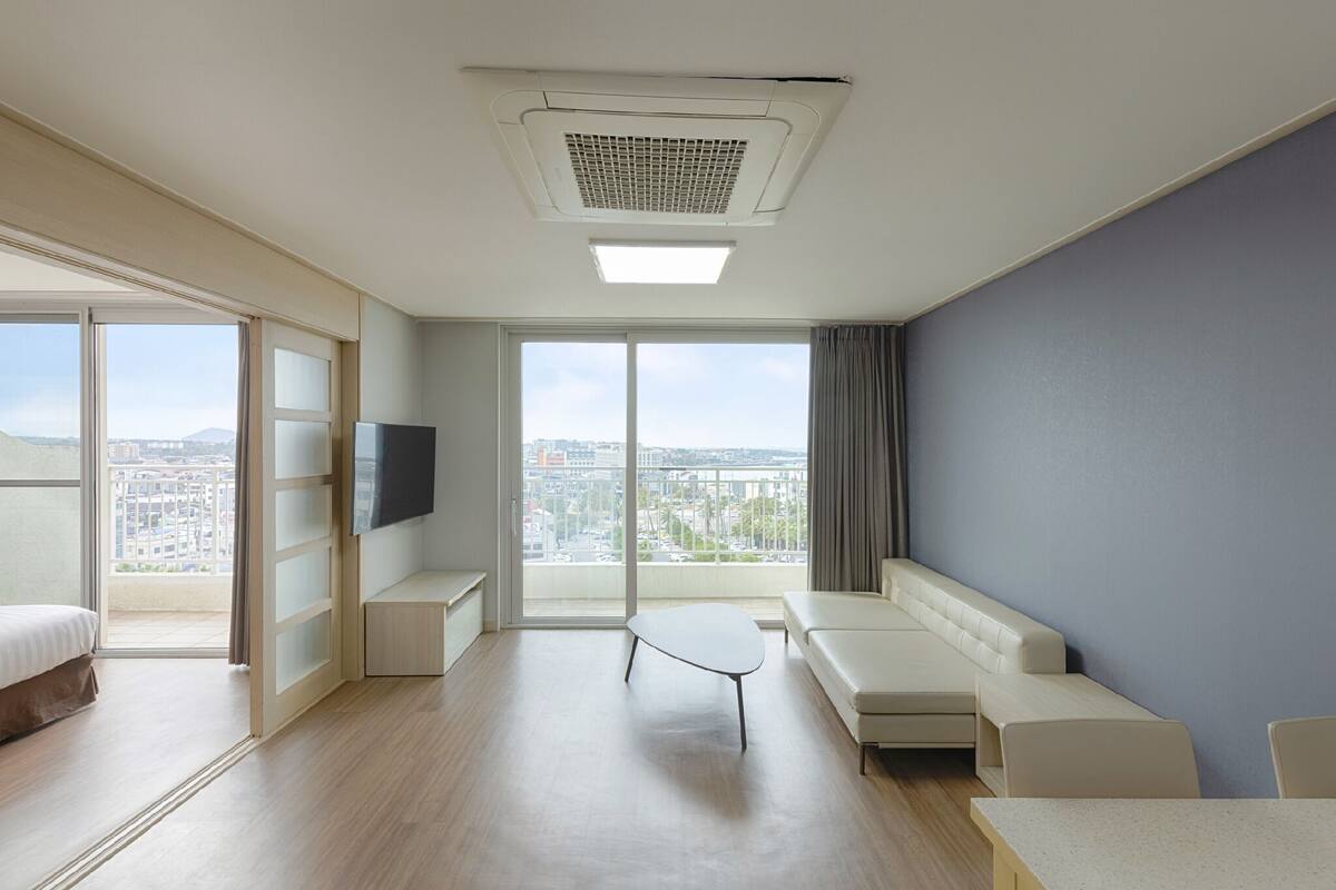 A modern hotel room with a large window, a flat-screen TV, a sofa, a small table, and a bed.